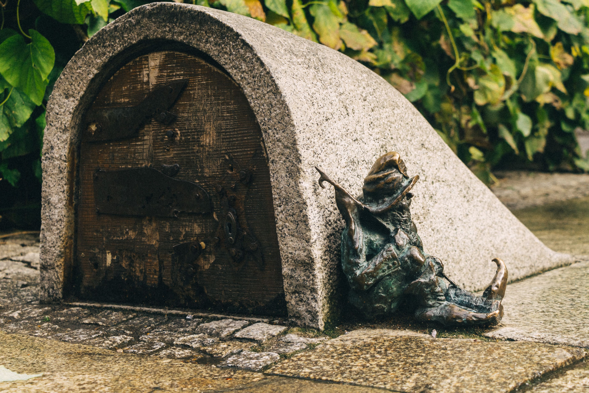 A small bronze Wrocław dwarf (krasnal) sitting beside a stone dwarf house with an iron door, ivy growing above it